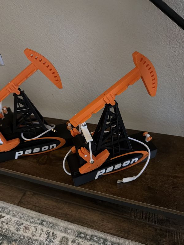 Two orange and black 3D-printed pumpjack desk models with a company logo on the base and a USB cable